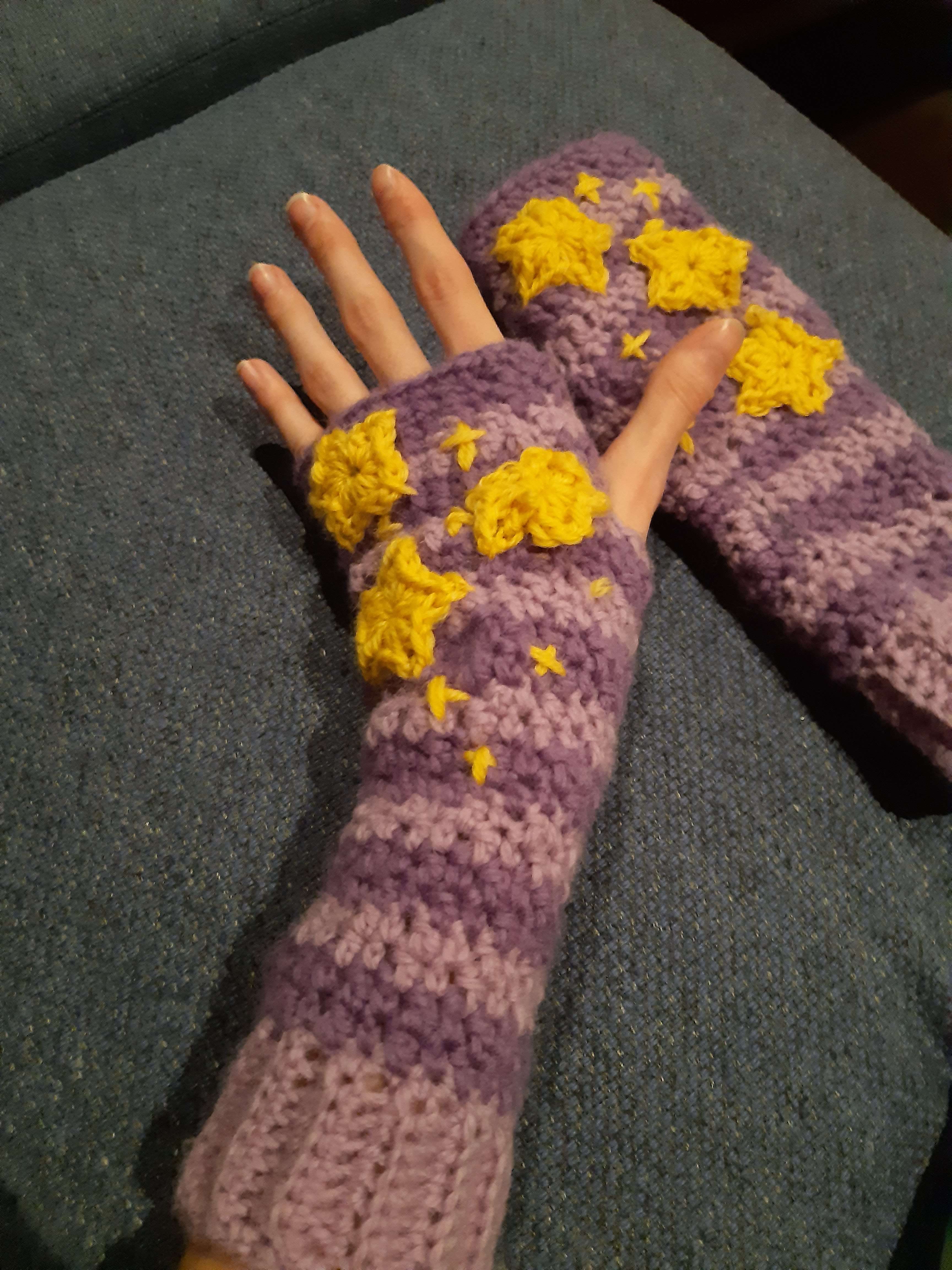 a pair of purple and lighter-purple striped crocheted handwarmers. they have yellow crocheted stars sown into them.